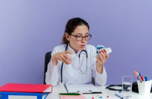 impressed young female doctor wearing medical robe and stethoscope sitting at desk with medical tools holding and looking at medical pills and holding beaker isolated on purple background